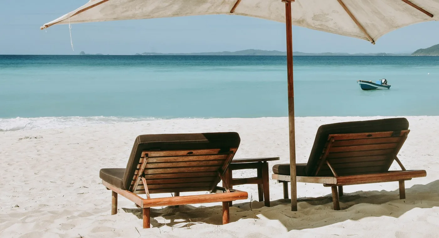 Two wooden beach loungers under a white umbrella by turquoise water